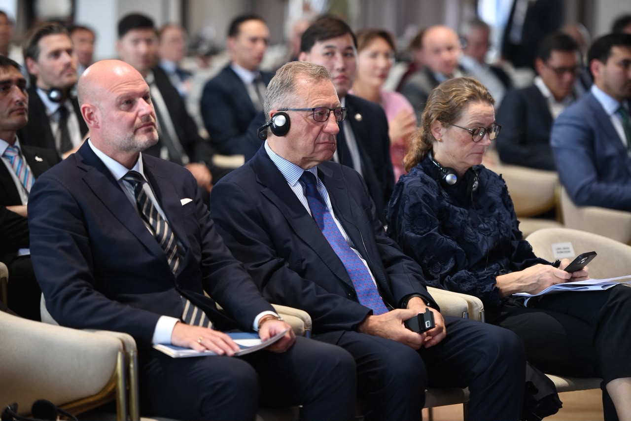 Ресми киімдегі адамдар жиналыста отыр, кейбіреулері құлаққап таққан.