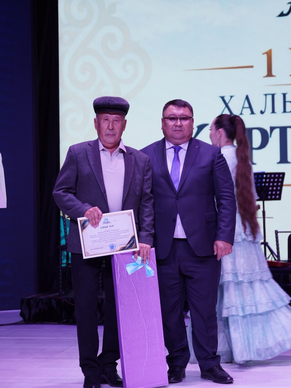 Сыйлық пен алғыс хатты ұстаған екі ер адамның сахнадағы суреті.