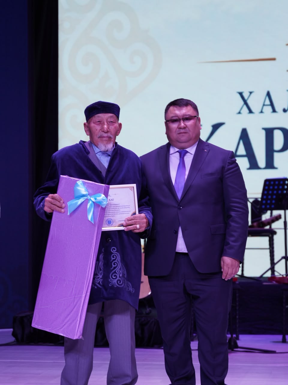 Сыйлық пен грамота алған қазақ киіміндегі қарт адам және костюм киген ер адам.