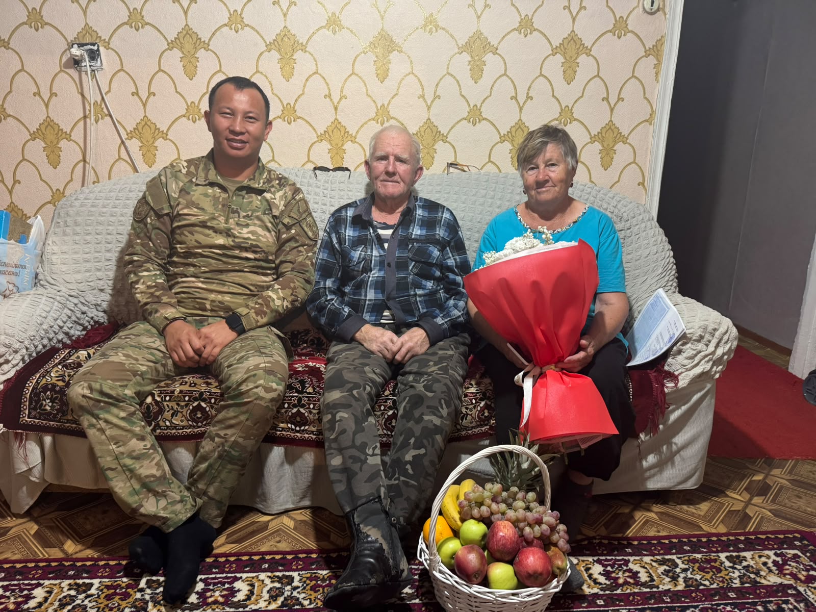 Мужчина в военной форме, пожилой мужчина и женщина с букетом цветов сидят на диване в комнате. Перед ними корзина с фруктами.