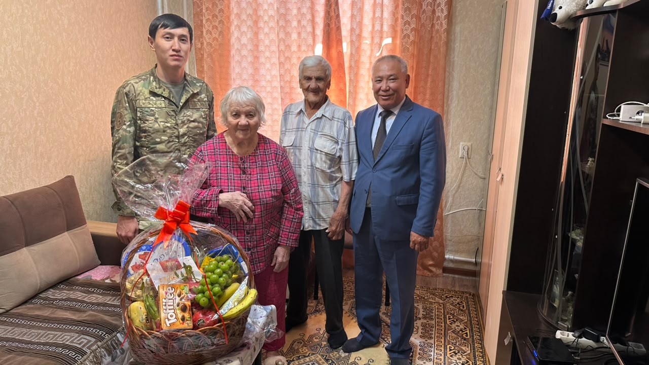 Группа из четырех человек: мужчина в военной форме, пожилая женщина, пожилой мужчина и мужчина в синем костюме с корзиной подарков.
