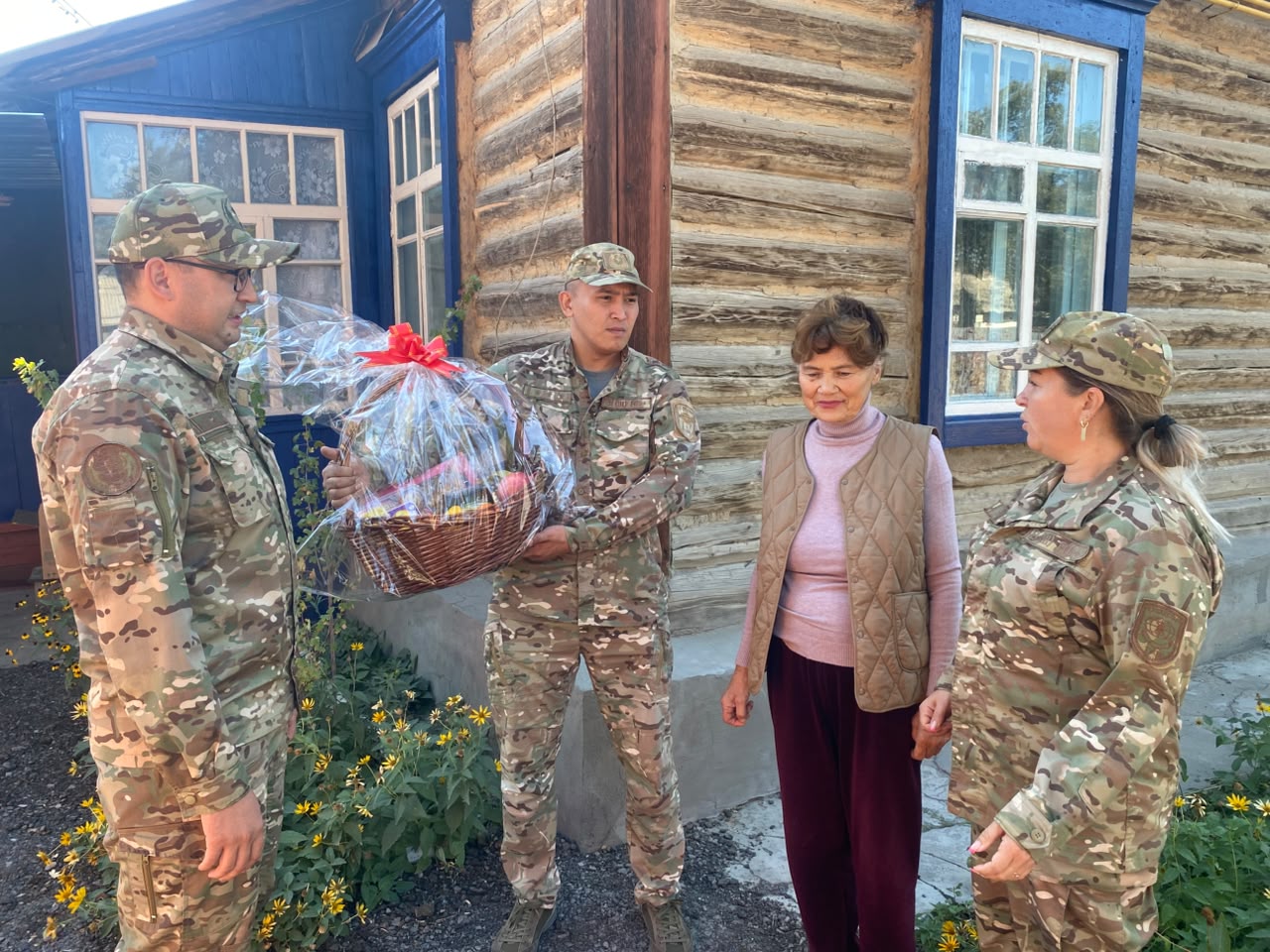 Военнослужащие в камуфляже вручают подарочную корзину женщине на фоне деревянного дома.
