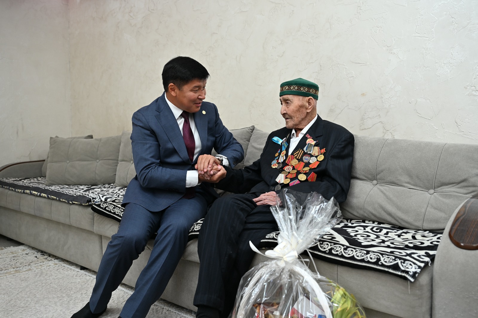 Костюм киген жас ер адамның медальдар таққан қарт адаммен сөйлесіп, қолынан ұстап отырғаны.