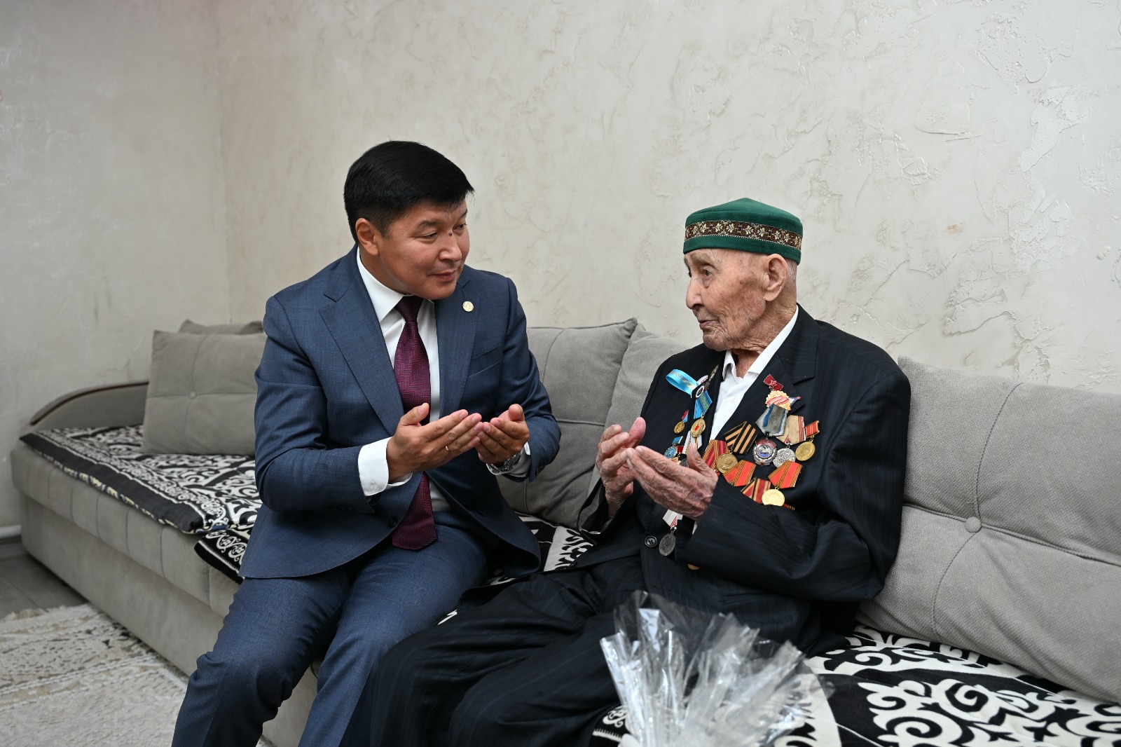 Костюм киген жас ер адам мен жүзінде медальдары бар қарт кісінің әңгімелесуі.