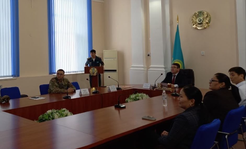 Жиналыс залындағы ресми кездесу: трибунада сөйлеп тұрған әскери қызметкер және үстел басындағы қатысушылар.
