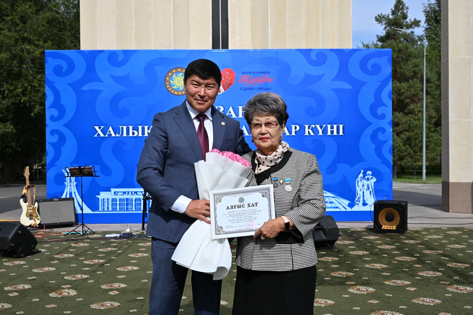 Костюм киген ер адам әйел адамға гүл шоғы мен алғыс хат табыстап жатыр.