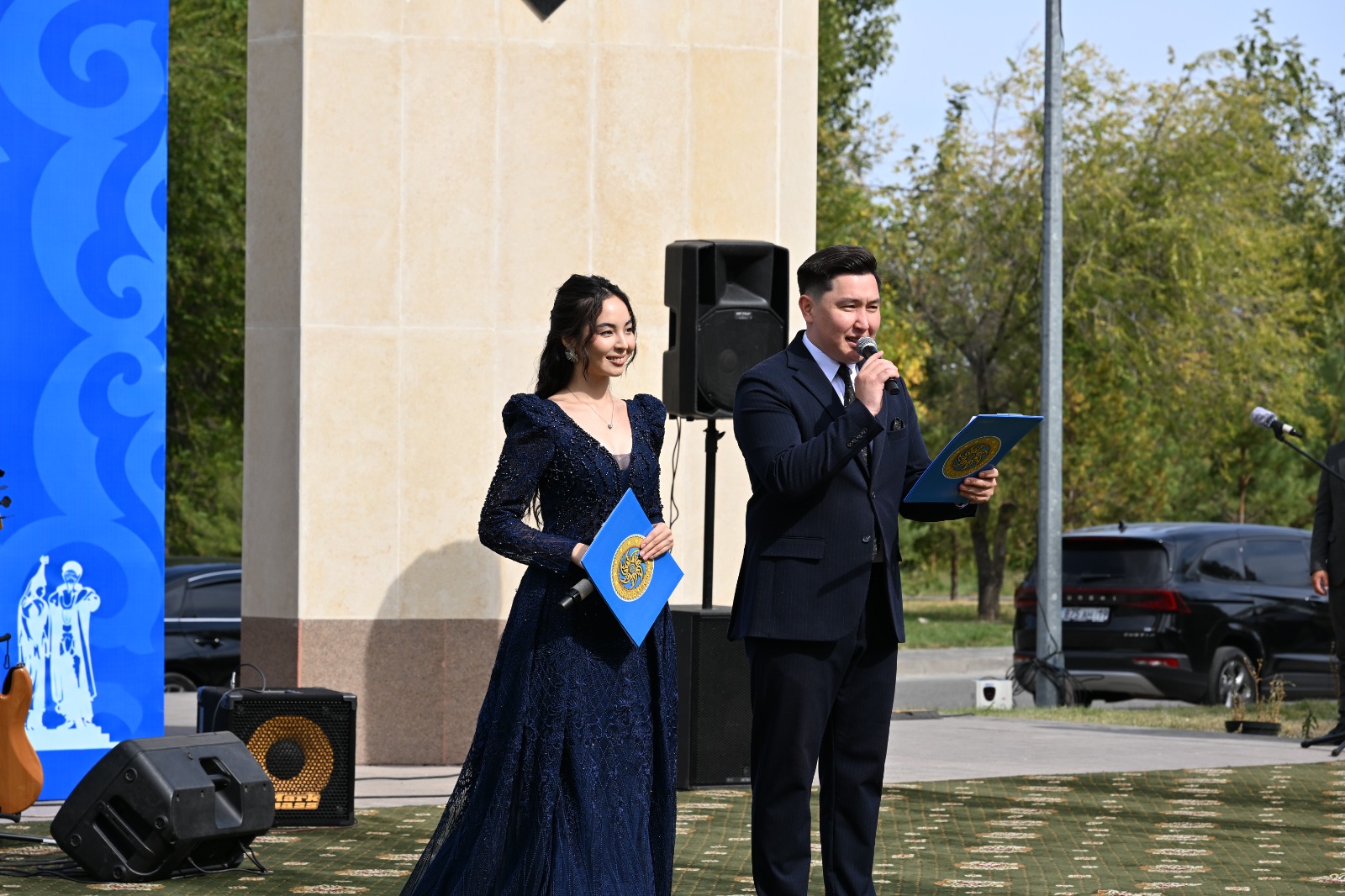 Көк көйлек киген қыз бен костюм киген жігіт сахнада микрофонмен сөйлеп тұр.