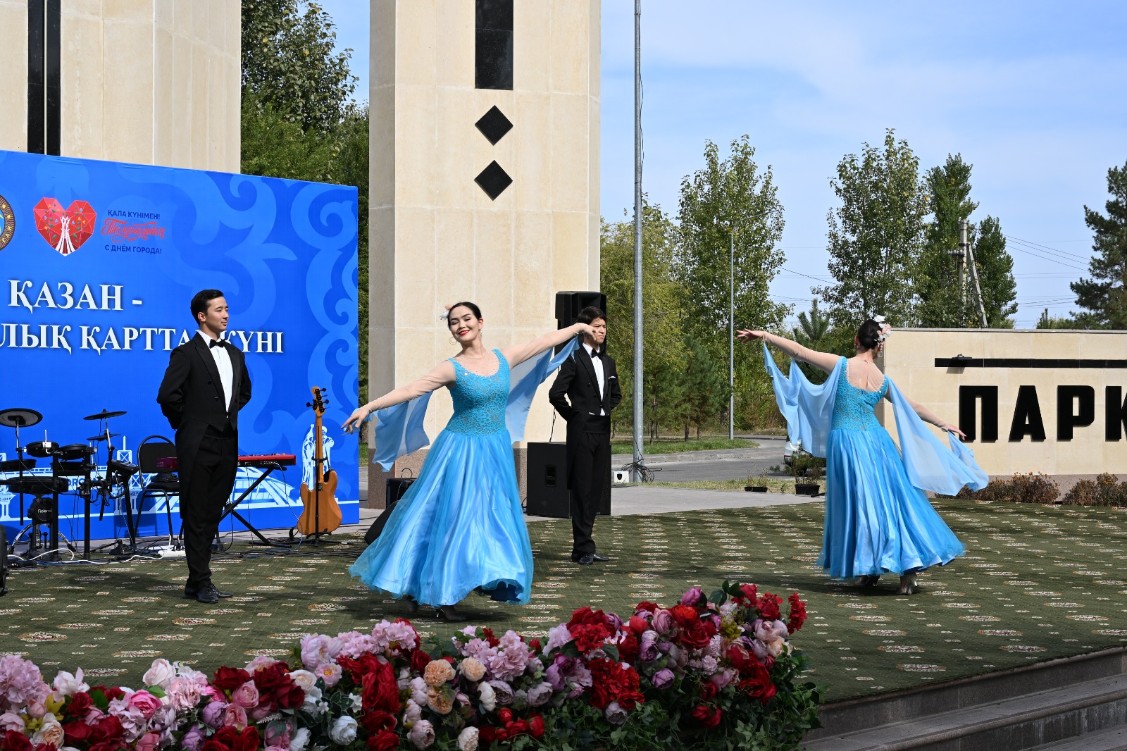 Көк көйлек киген бишілер мен смокинг киген ер адамдардың ашық алаңдағы қойылымы.