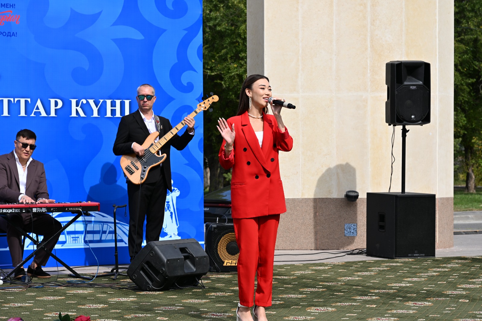 Қызыл костюм киген әнші мен музыкалық топтың ашық алаңдағы қойылымы.