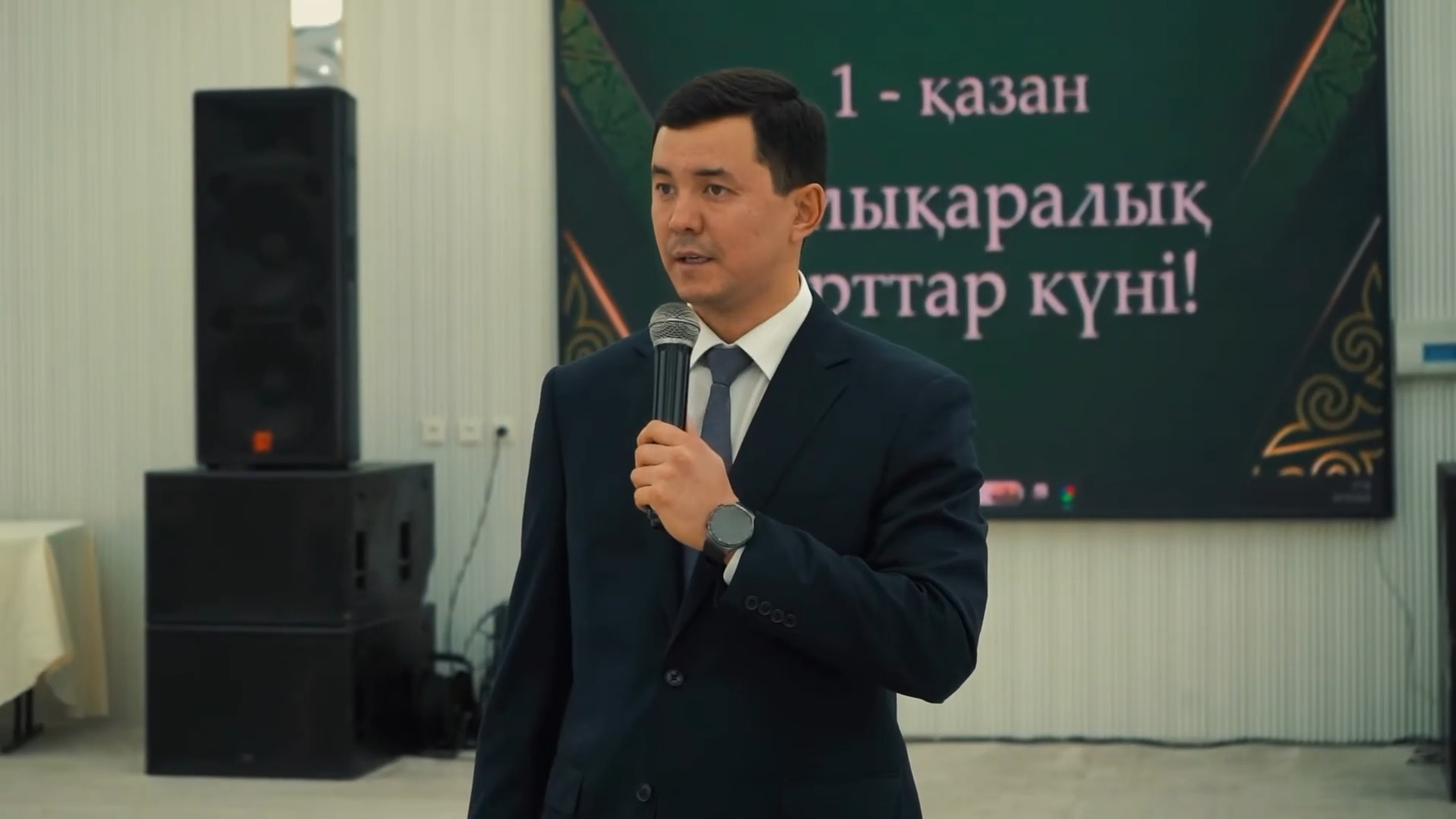 Костюм киген ер адам микрофонмен сөйлеп тұр, артында «1-қазан» деп жазылған экран.