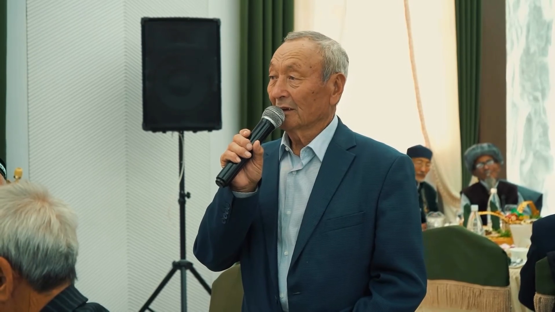 Микрофонмен сөйлеп тұрған костюм киген қарт адам.