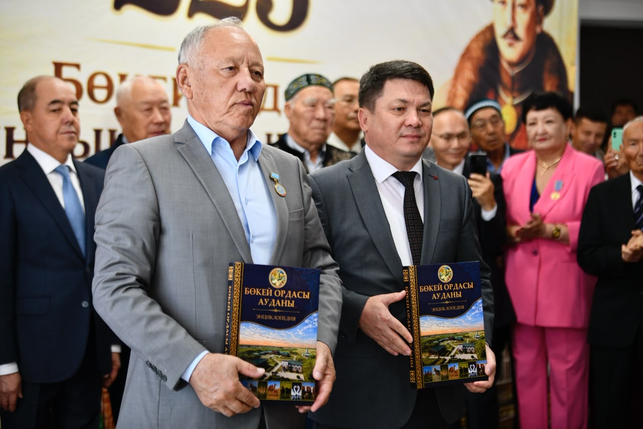 Двое мужчин в костюмах держат в руках книги с надписью «Бөкей ордасы ауданы энциклопедия».