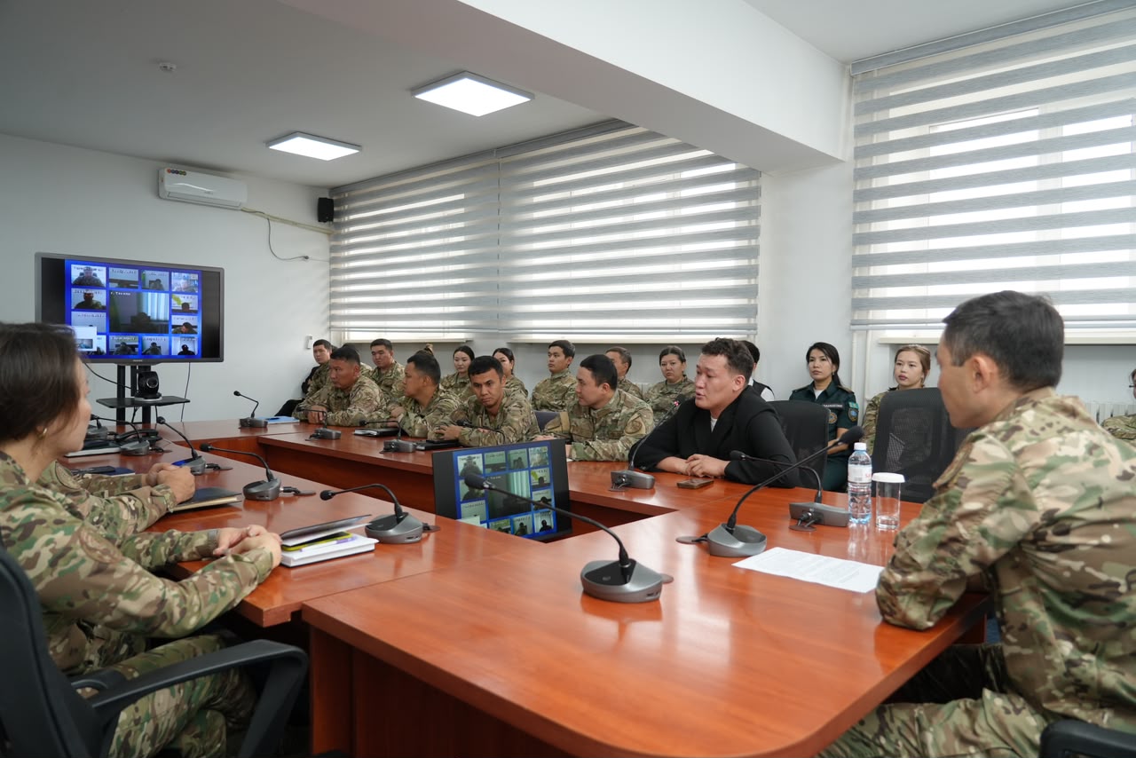 Военные в камуфляже и гражданские на совещании в конференц-зале с мониторами и системой видеосвязи.