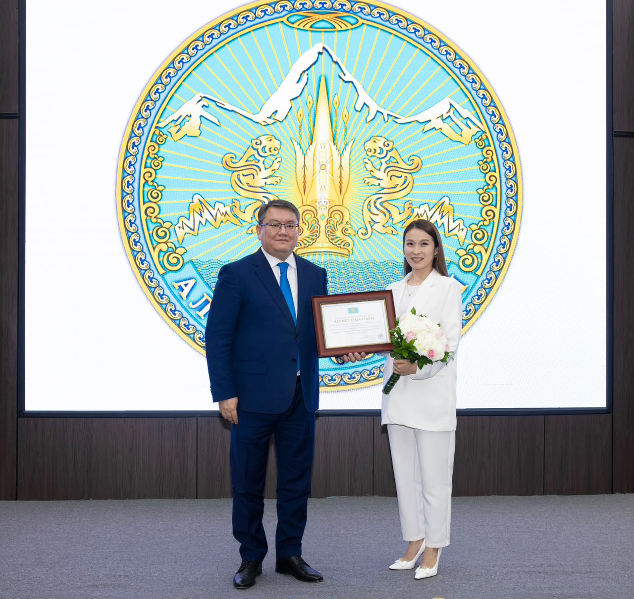 Көк костюмді ер адам мен ақ костюмді әйел сыйлықаты мен гүл ұстап, үлкен эмблеманың алдында тұр.