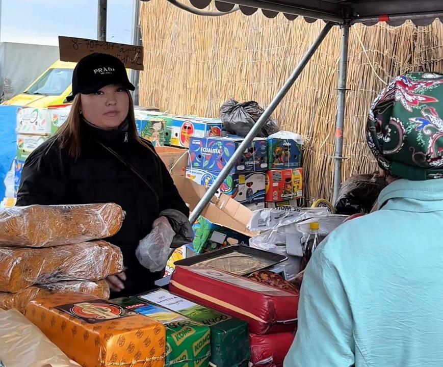 Базардағы сауда нүктесінде тұрған сатушы әйел және алдындағы тағамдар мен тауарлар.