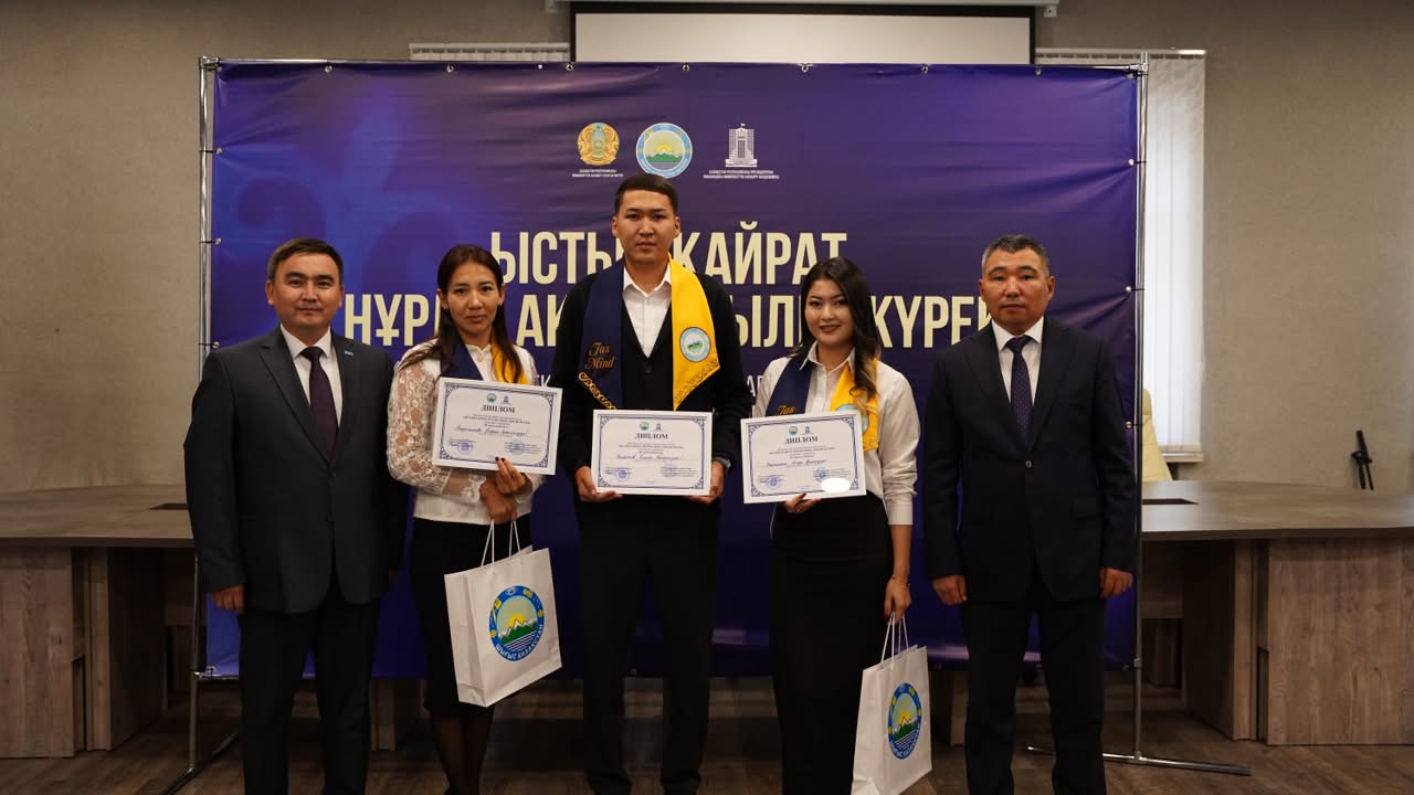 Диплом алған үш студент пен екі ресми киінген ер адамның көк фондық баннер алдындағы суреті.