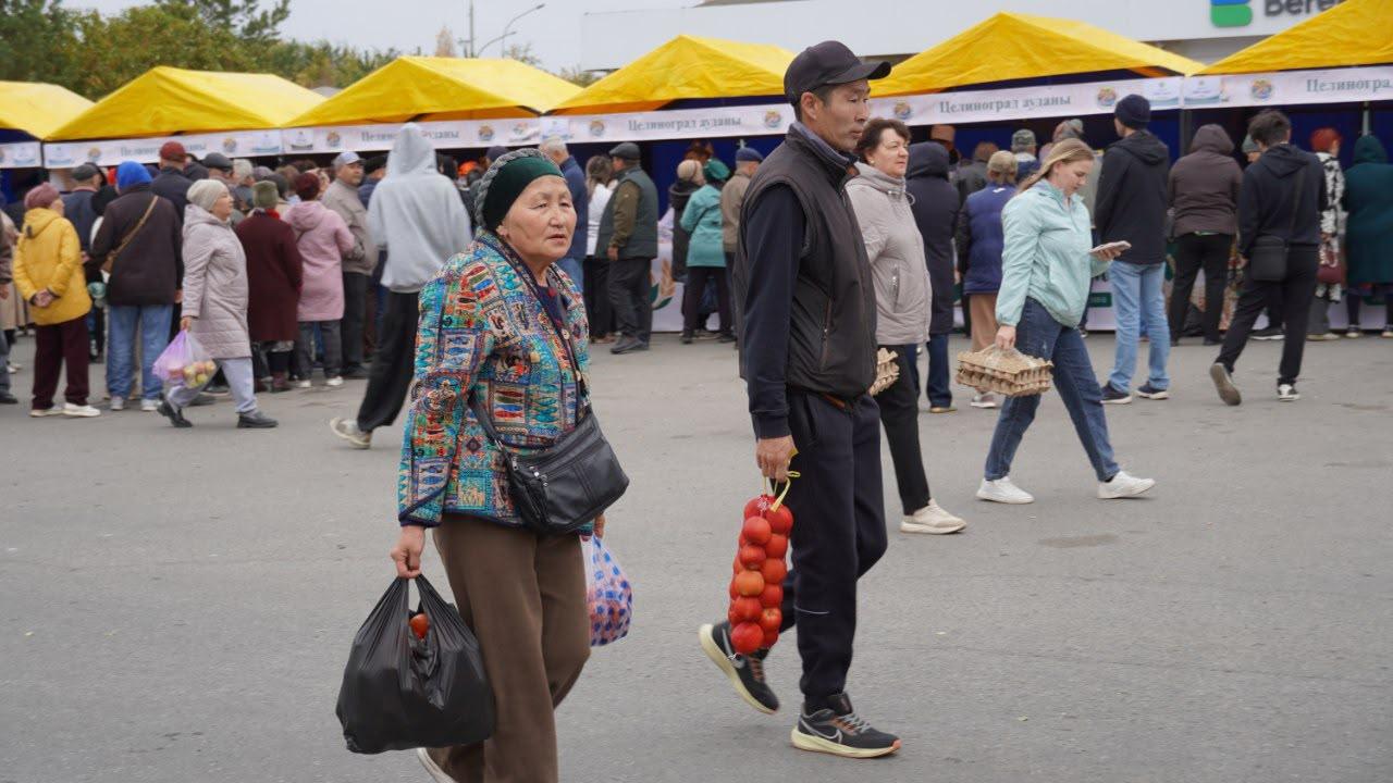 Люди на открытом рынке под желтыми тентами; на переднем плане женщина с сумками и мужчина с нитью помидоров.