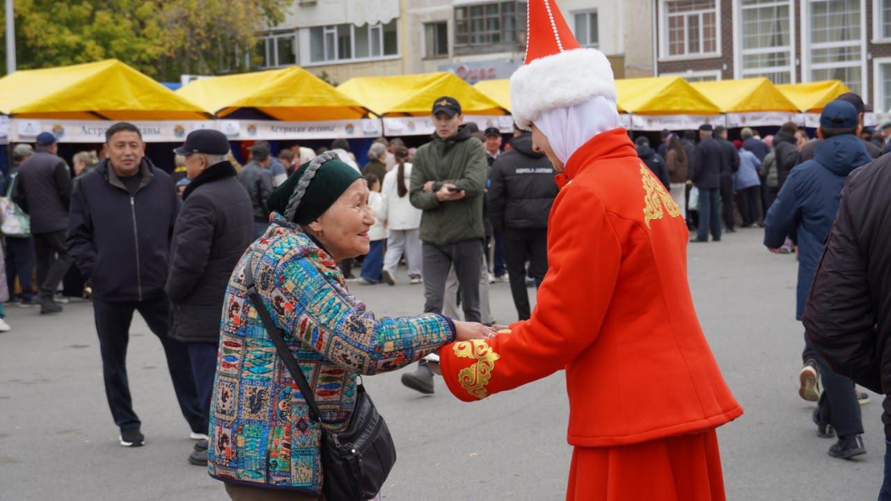 Пожилая женщина в цветной куртке пожимает руку девушке в красном костюме Снегурочки на городской площади.