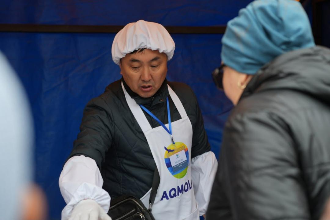 Мужчина в белом колпаке, фартуке с надписью «AQMOLA» и темной куртке разговаривает с женщиной на фоне синего тента.
