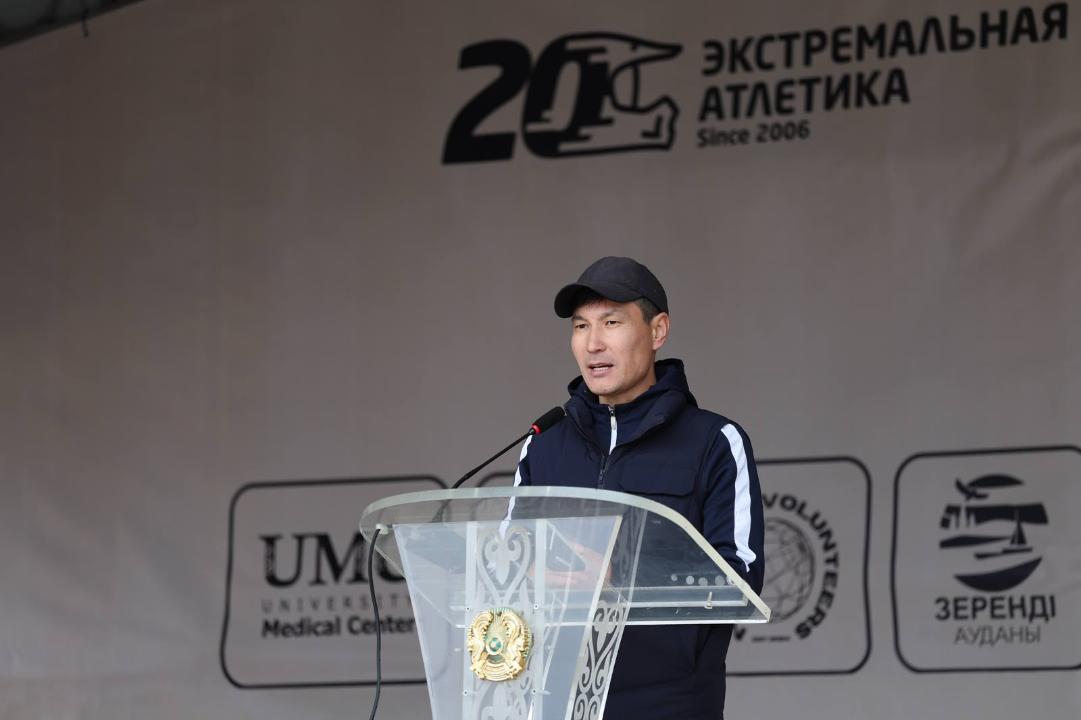 Мужчина в темной кепке и куртке выступает с речью за прозрачной трибуной на фоне баннера