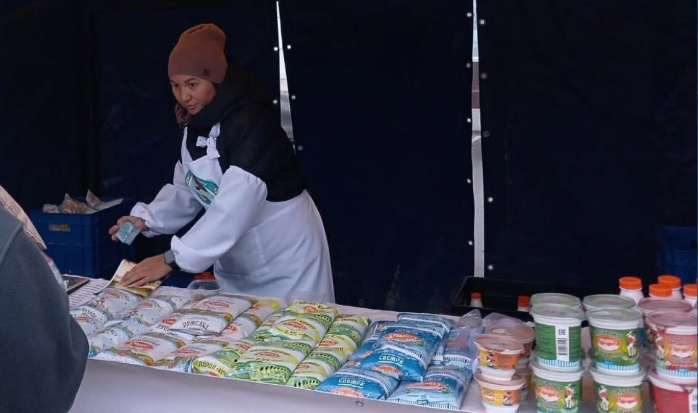 Женщина в белом фартуке и коричневой шапке продает молочные продукты на рынке.