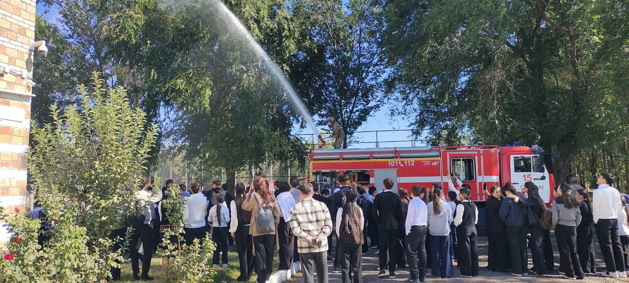 Өрт сөндіру көлігі мен өрт сөндірушілердің жұмысын бақылап тұрған оқушылар тобы.