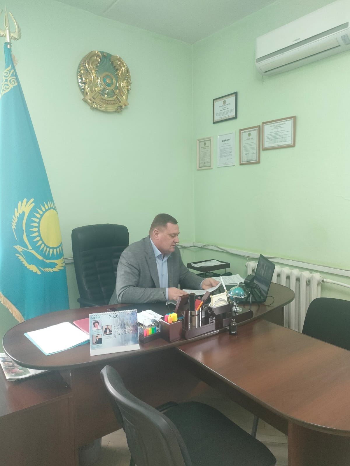 Мужчина в сером пиджаке сидит за рабочим столом в кабинете с флагом и гербом Казахстана на стене.