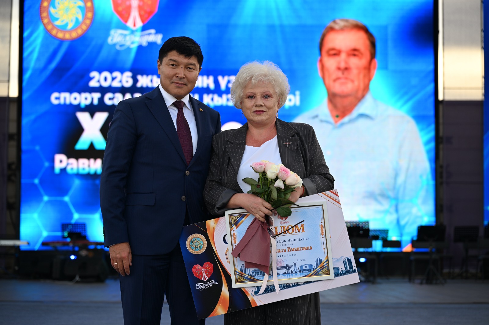 Сыйлық пен гүл ұстаған ер адам мен әйелдің сахнадағы суреті.