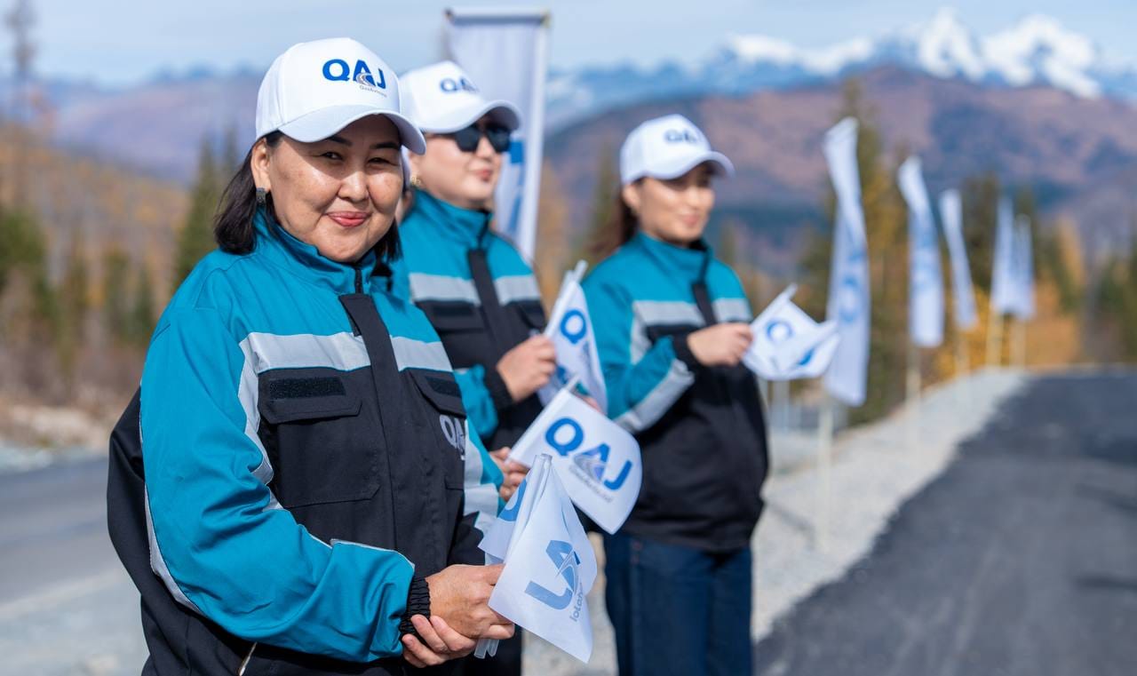 QAJ логотипі бар форма киген үш әйел жол жиегінде тұр.