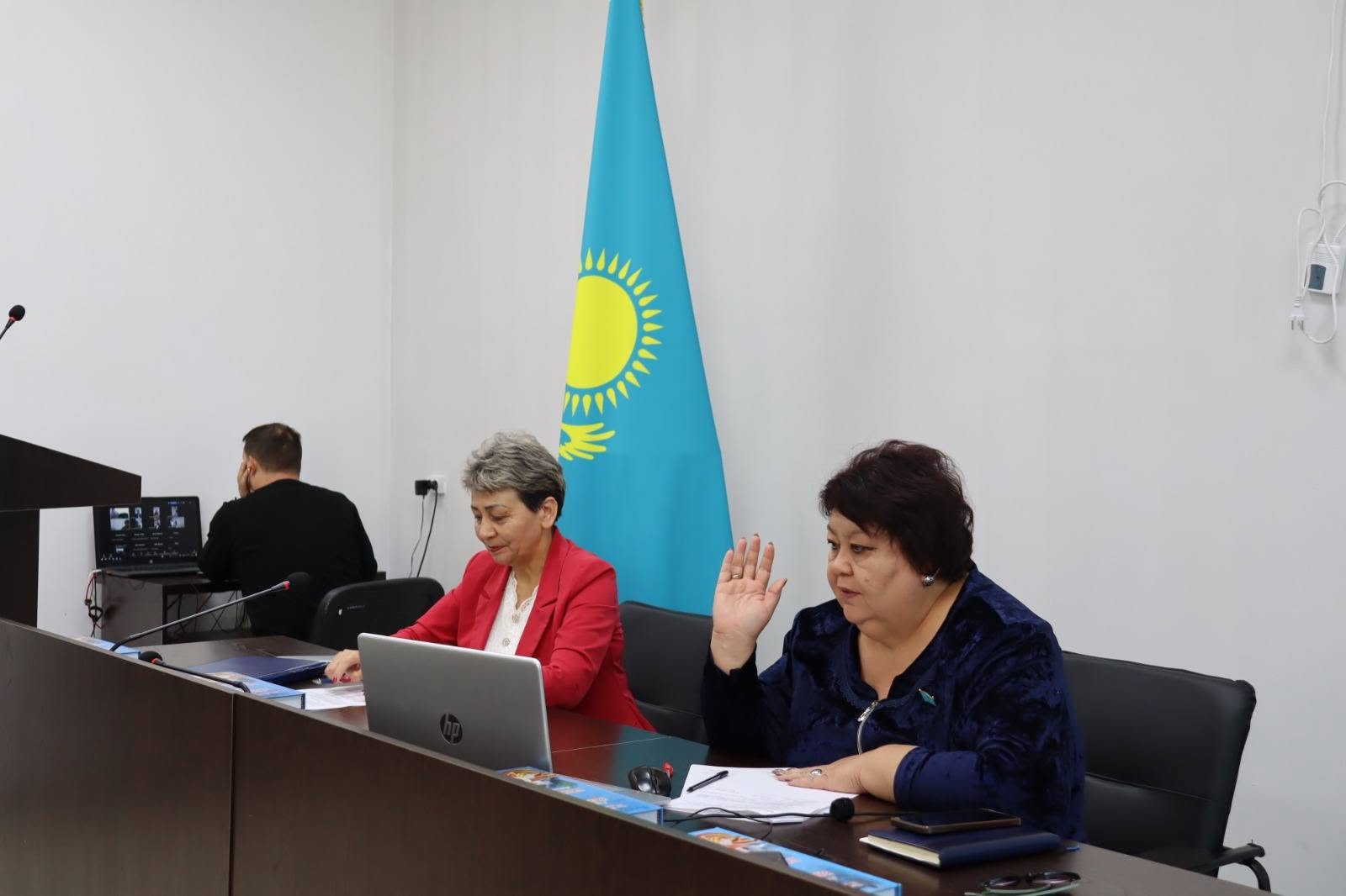 Қазақстан туының алдында ноутбукпен жұмыс істеп отырған екі әйел және артқы жақтағы ер адам.