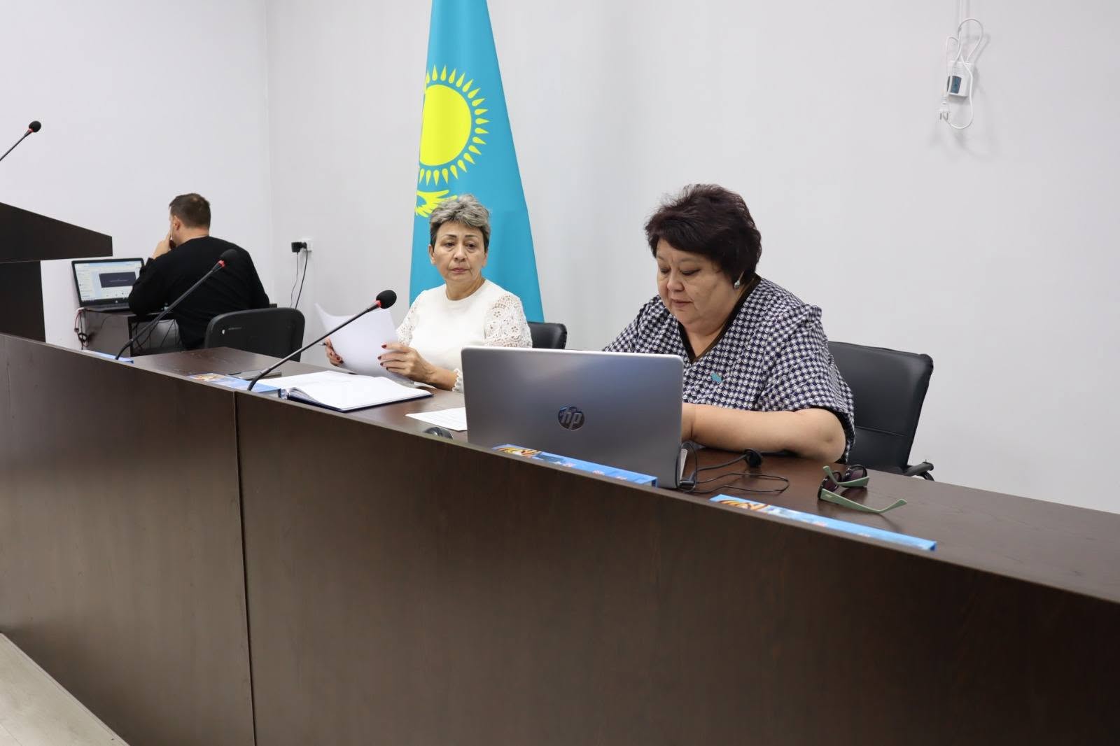 Қазақстан туының алдында жиналыс үстелінде отырған екі әйел және ноутбук.