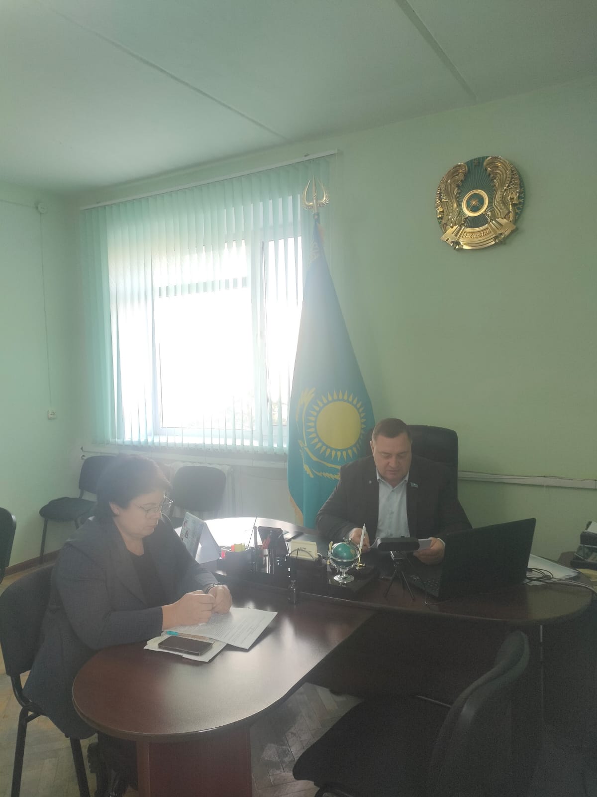 Кеңседе үстел басында жұмыс істеп отырған екі адам. Фонда Қазақстан Республикасының туы мен елтаңбасы бейнеленген.