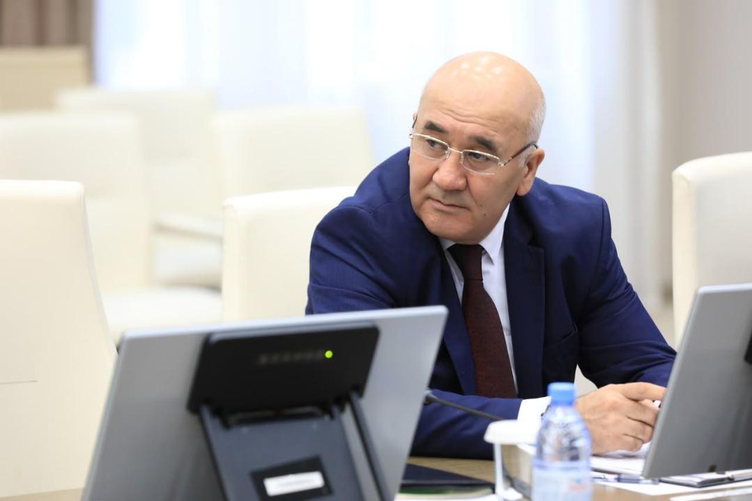 Көк костюм мен көзілдірік киген, ноутбук алдында отырған ер адам.