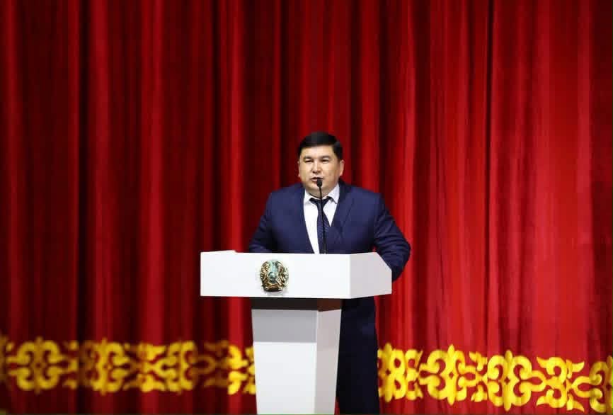 Қызыл перде алдындағы трибунада сөйлеп тұрған костюм киген ер адам.