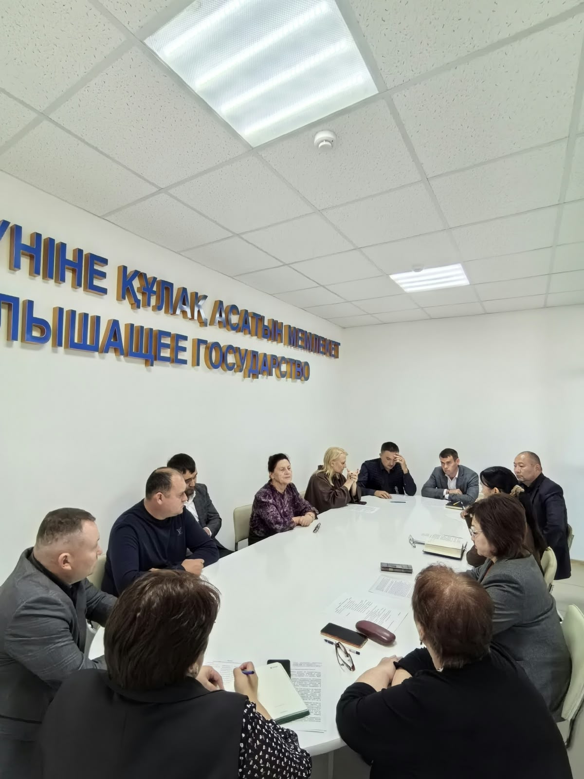 Кеңседегі мәжіліс үстелі төңірегінде жиналған адамдар тобы.