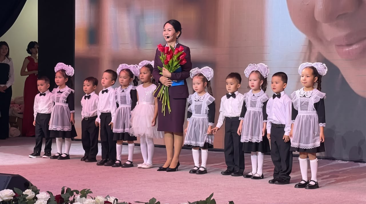 Сахнада гүл ұстаған әйел және киімдері үйлестірілген кішкентай балалар тобы.