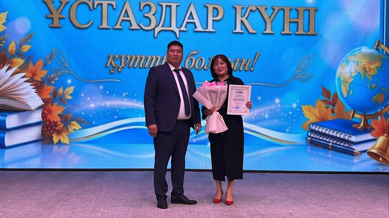 «Ұстаздар күні» жазуы бар экран алдында тұрған, қолында гүл мен грамотасы бар әйел мен костюм киген ер адам.