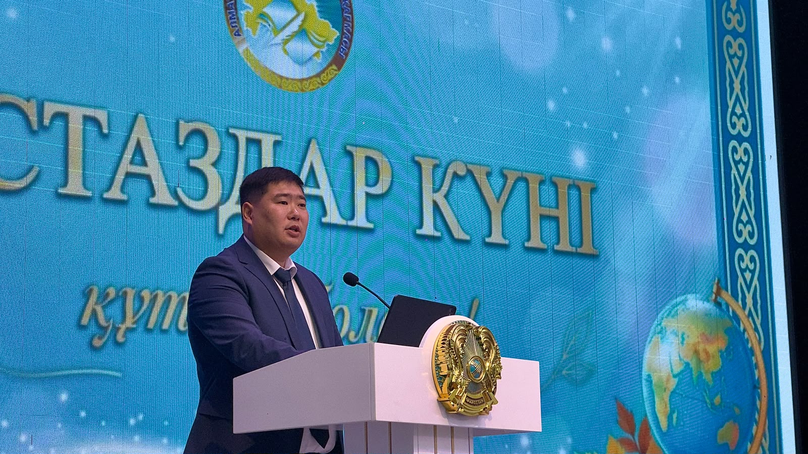 Трибунада сөйлеп тұрған ер адам. Артында «Ұстаздар күні» жазылған көк экран.