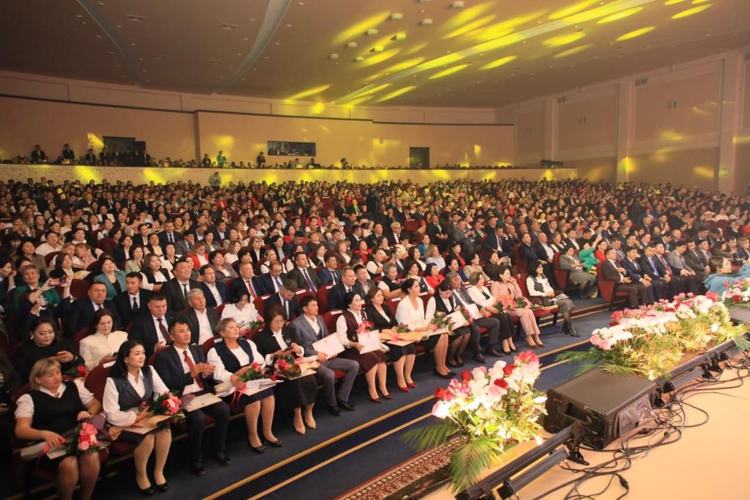 Жиында жиналған көптеген адамдардың залда отырған көрінісі.
