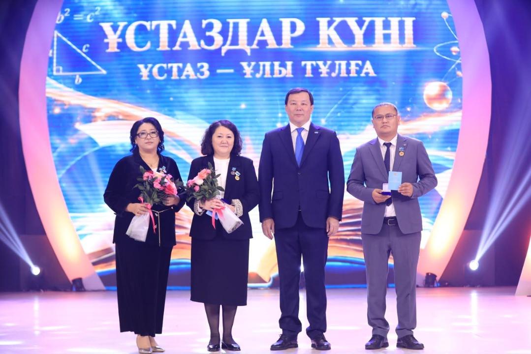 «Ұстаздар күні» мерекелік іс-шарасында сахнада тұрған төрт адам.