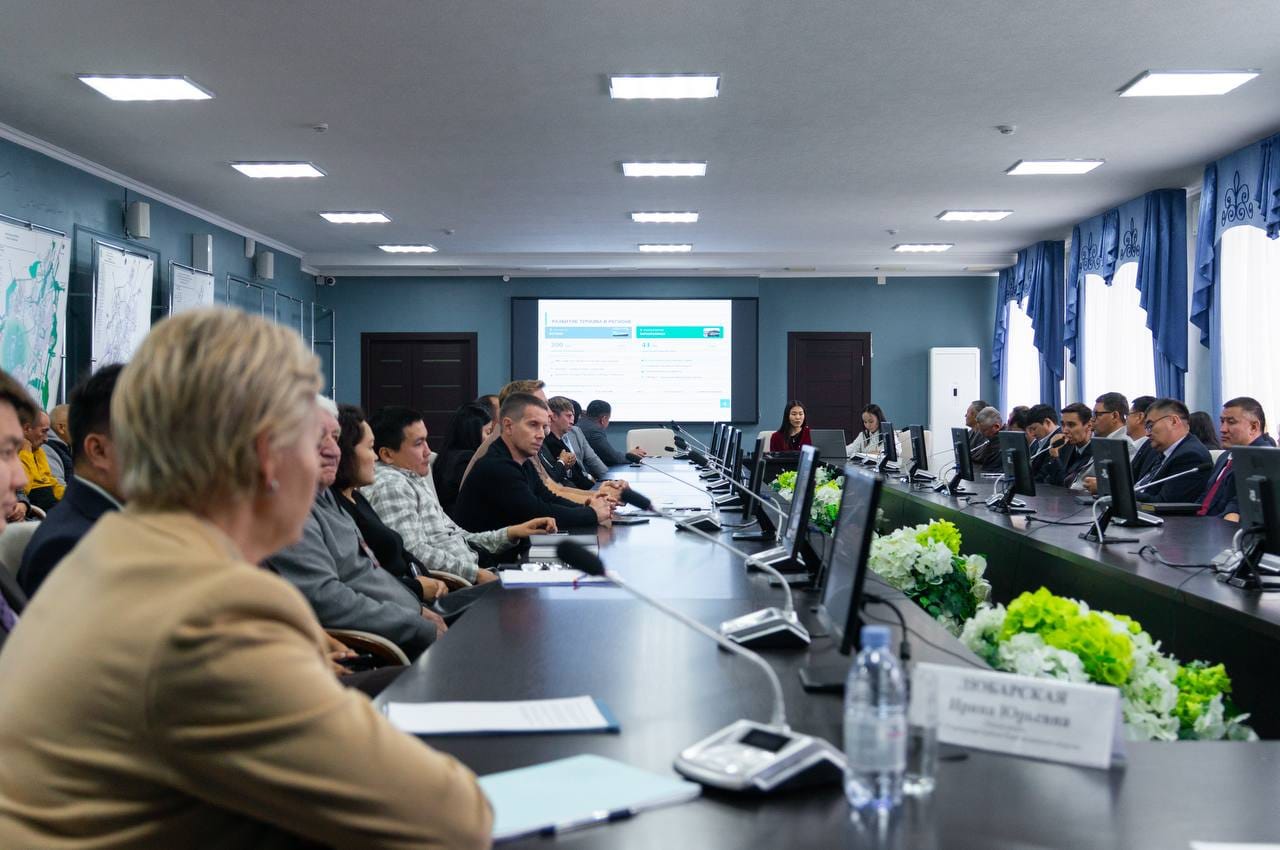 Жиналыс залындағы мәжіліс: үстел басында отырған қатысушылар және алдындағы презентация экраны.