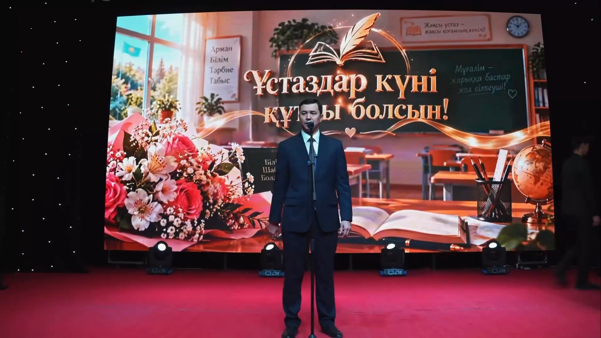 «Ұстаздар күні құтты болсын!» деп жазылған экран алдында сөйлеп тұрған костюм киген ер адам.