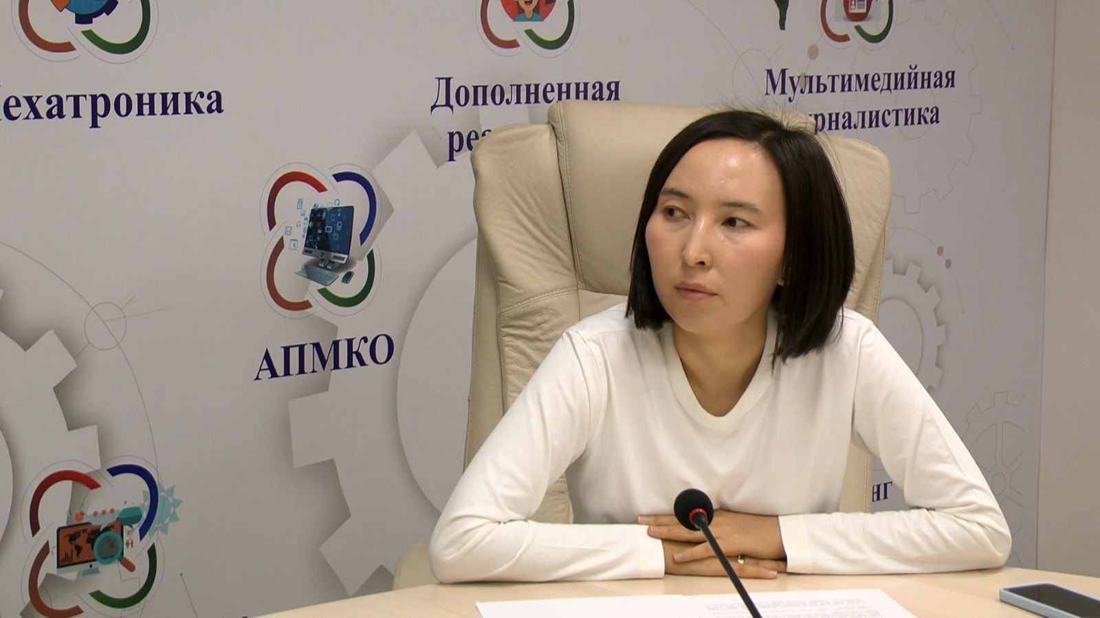Ақ киімді әйел адам микрофонның алдында отыр, артында «АПМКО» жазуы бар баннер тұр.