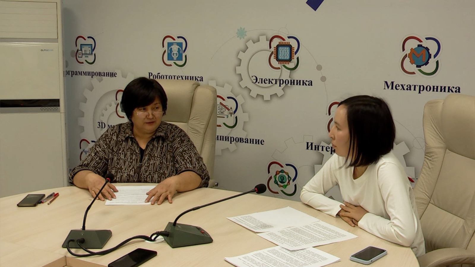 Келіссөз жүргізіп жатқан екі әйел. Фонда робототехника мен электроника туралы жазулар бар.