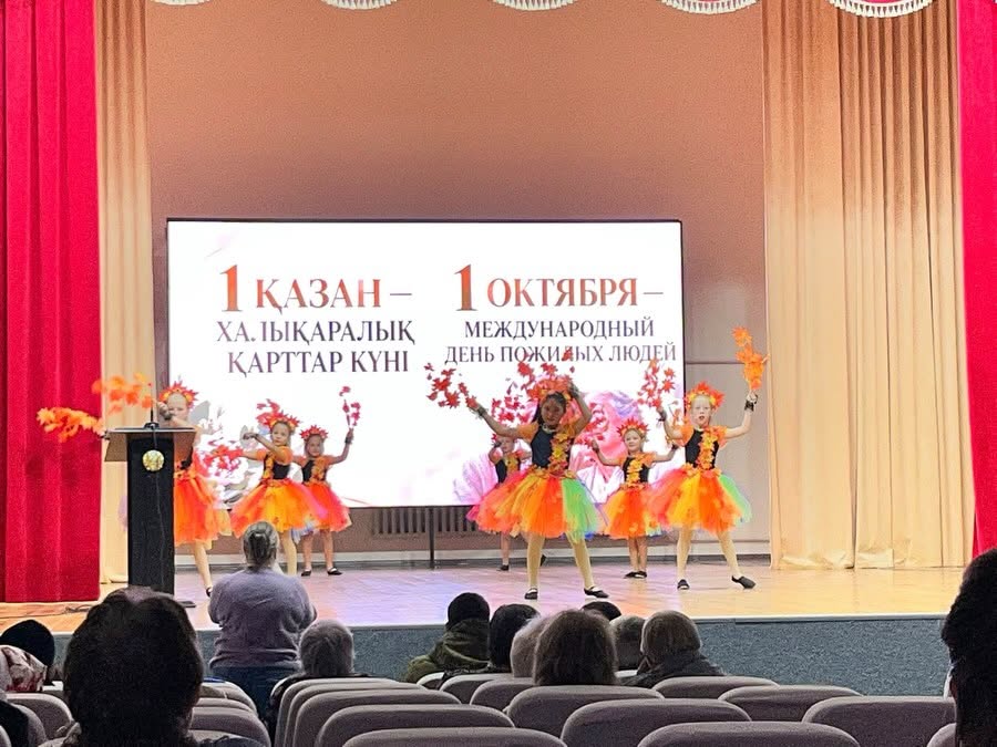 Қызғылт сары түсті костюм киген балалар сахнада «Халықаралық қарттар күні» деген жазу көрсетіліп тұрған экран аясында би билеп жатыр