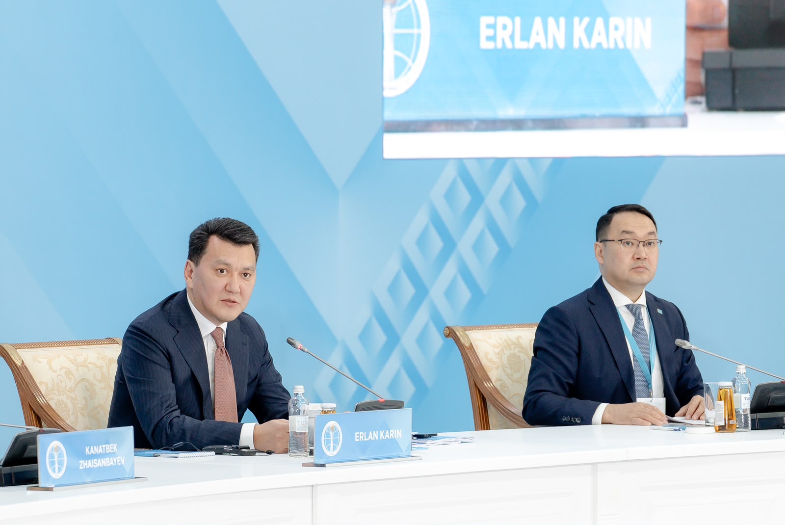 Кіссада екі ер адам костюм киіп, конференция үстелінде отыр. Артында «ERLAN KARIN» жазуы бар экран көрінеді.