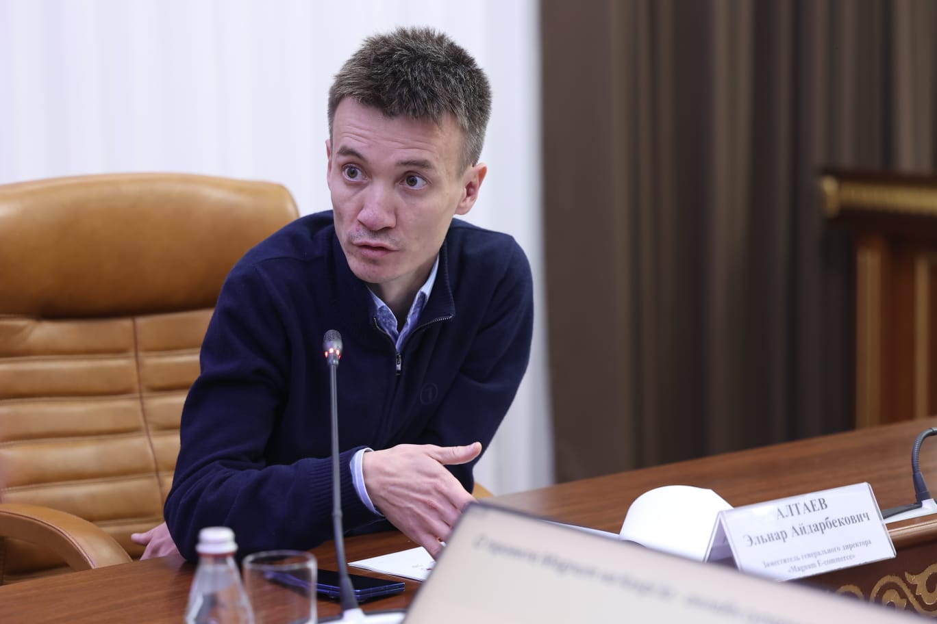 Көк түсті киім киген ер адам жиналыста сөйлеп тұр.