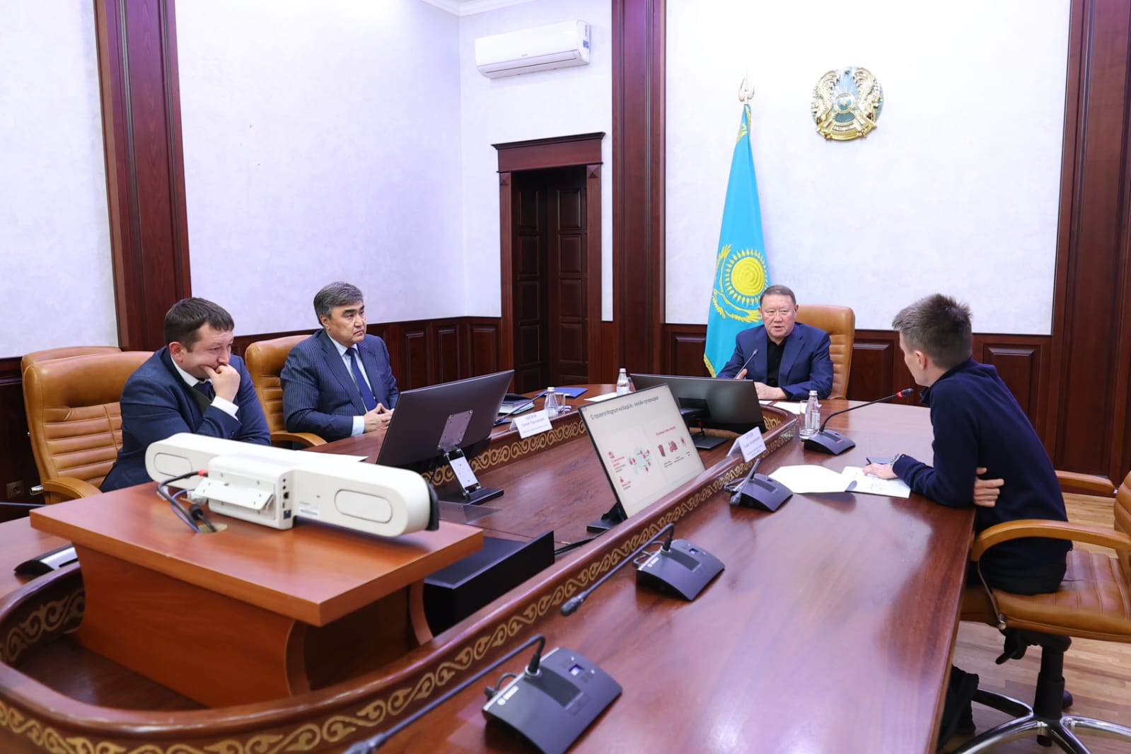 Қазақстан Республикасының мемлекеттік рәміздері бар бөлмедегі ресми жиналыс. Үстел басында төрт ер адам отыр.