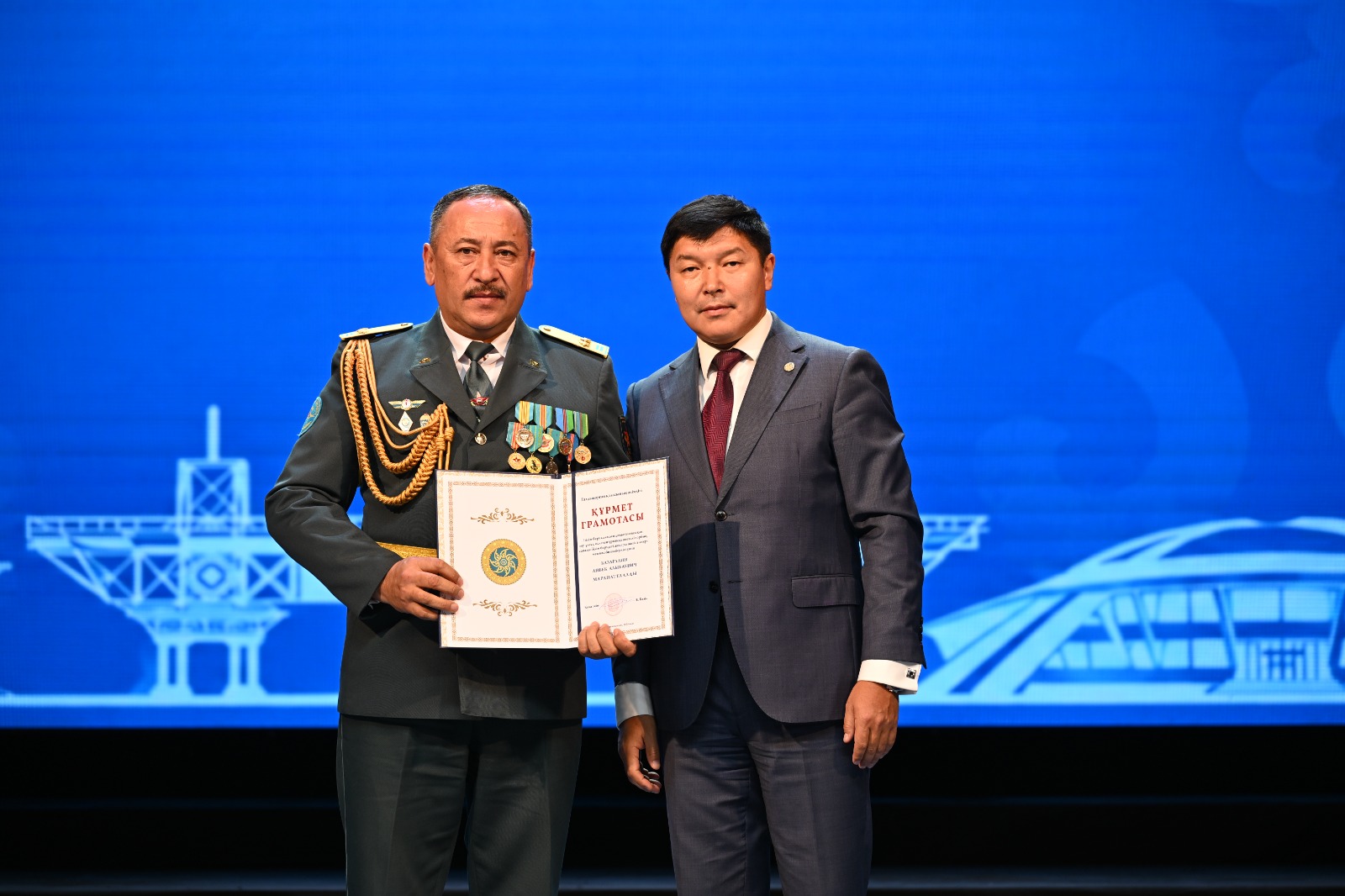 Әскери формадағы ер адам мен костюм киген ер адамның қолға сертификат ұстап тұрған суреті.