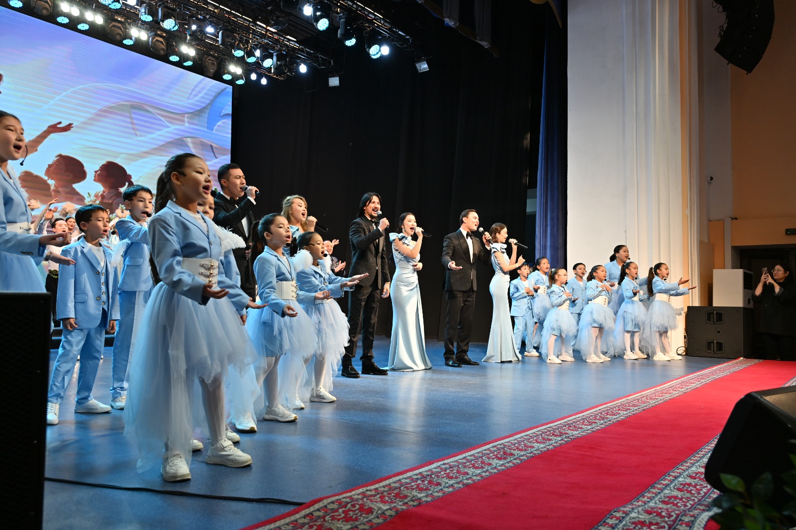 Сахнада көк киімді балалар мен костюм киген ересектердің хоры ән айтып тұр.